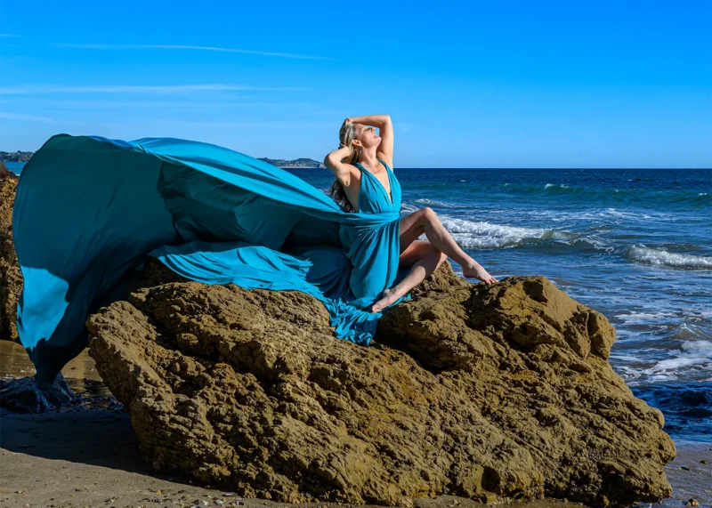 Malibu Beach Flying Dress Experience Photoshoot with beautiful woman in a blue dress sitting on the rocks on the beach