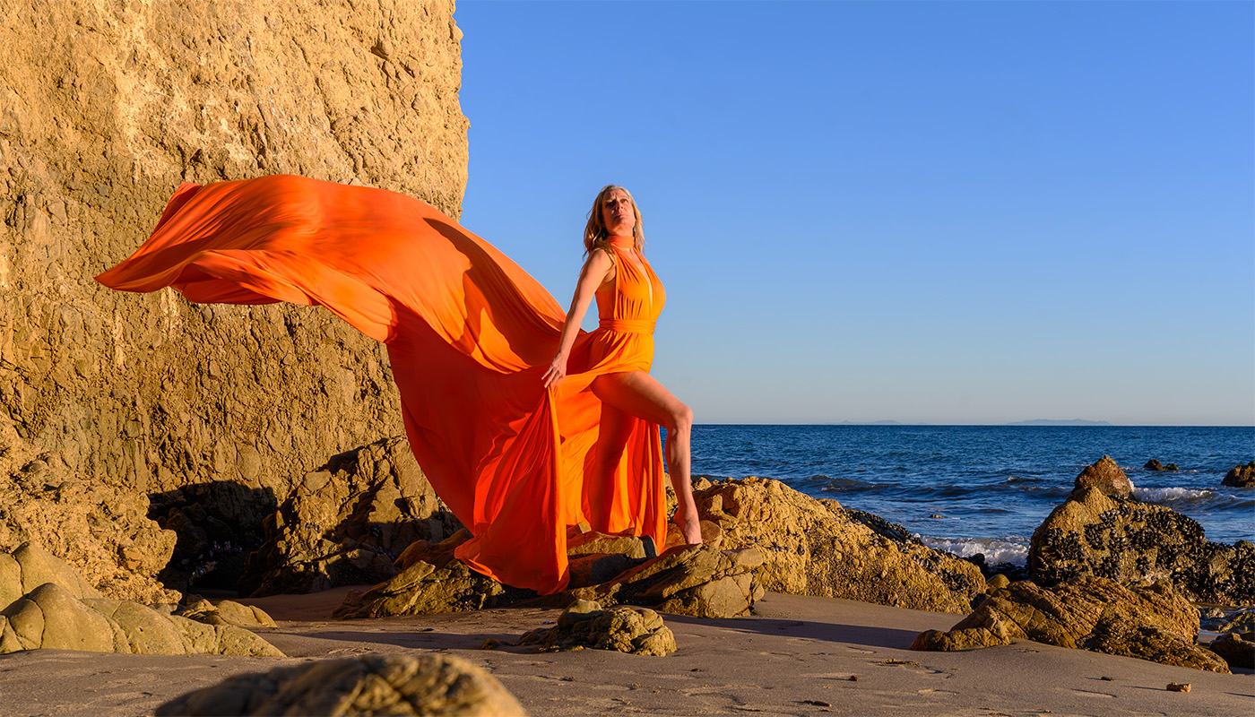 Middle-aged woman standing on rocks on the Californian coast in a bright orange flying dress